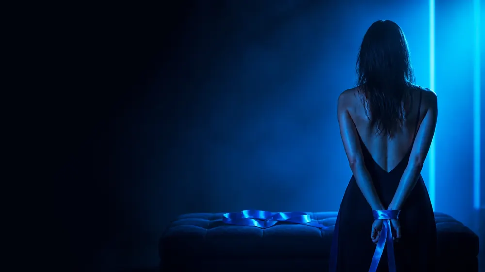 OnlyFans tvůrci v nice Bare Bottom Spanking — žena s hedvábnou stuhou kolem zápěstí v neonovém světle, sametová lavička a poskládaná hedvábná stuha čekající
