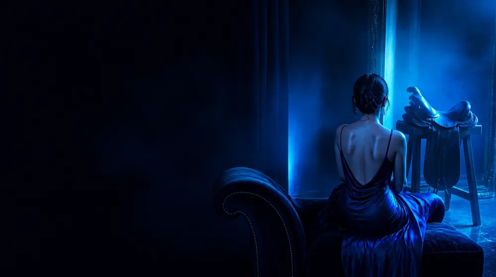 Reverse Cowgirl OnlyFans creators — woman in a silk slip dress reclining on a chaise longue, a saddle stand facing away from the mirror