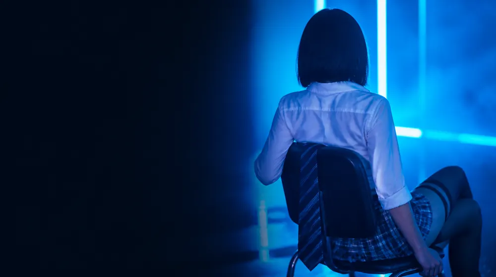 Schoolgirl OnlyFans creators — woman in a plaid skirt in a neon-lit room, a tie loosened over the chair back